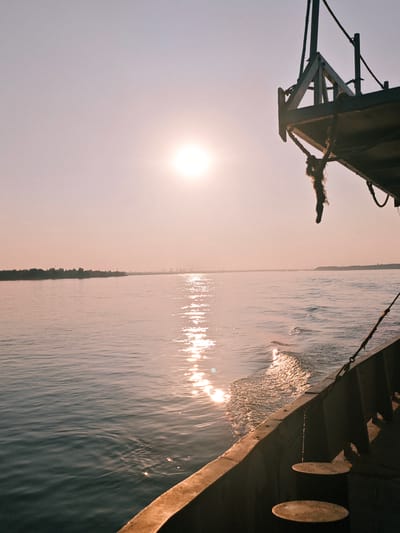 The image depicts a wide, calm body of water, likely a river, viewed from the deck of a boat. The sun is low in the sky, casting a warm, soft glow and creating a shimmering reflection on the water's surface. In the distance, the hazy outline of a treelined shore is visible, punctuated by industrial structures like cranes and possibly buildings, suggesting a port or industrial area. The foreground is dominated by the side of the boat, showing a railing and some vertical wooden structures. The sky is a pale, hazy pinkish-orange. Without specific landmarks or textual information within the image, it is impossible to determine the precise location within the city of Lesosibirsk, Russia.