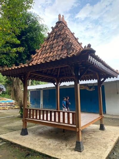 The image shows a traditional wooden gazebo with a tiled roof. The gazebo is located in Kecamatan Manyaran, Indonesia, at the coordinates -7.826 latitude and 110.816 longitude. The gazebo is situated in front of a building with blue doors. Two young boys are standing in front of the gazebo, one is facing the camera while the other is turned away. The gazebo is surrounded by foliage and is located in a rural or suburban area. The scene is captured on a cloudy day. The image is taken from an eye-level perspective. The gazebo is made of dark wood, and the tile roof is red-brown. The boys are wearing casual clothing. The building behind the gazebo is white with blue doors. The doors are closed. The environment is tranquil, and the scene is peaceful.