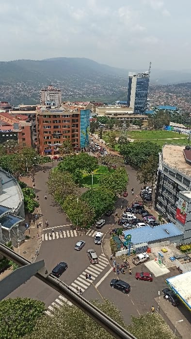 The image displays an aerial view of the Kigali City Roundabout (also known as Centenary Park Roundabout) in the central business district of Kigali City, Rwanda, observed during daytime under a partly cloudy sky.
The central feature is a large, multi-lane traffic roundabout with an irregular-shaped central island densely covered in mature green trees. Surrounding the roundabout are various multi-story buildings. To the north-east, the prominent Kigali City Tower, a tall, slender building with a blue glass facade and a distinctive curved top, is clearly visible. Other notable buildings include a multi-story brown building with numerous windows to the west of the roundabout, and a building with a grey/white textured facade situated immediately east. A petrol station, identifiable by its fuel pumps and branding (likely Engen), is located on the southeastern edge of the roundabout, actively serving vehicles.
The roads are busy with vehicular traffic, consisting of numerous passenger cars (sedans and SUVs) in various colors including black, silver, white, and red. Several motorcycles, locally known as boda-bodas, are also present, some individually, others in small clusters. Traffic flow appears active, with some vehicles stationary at intersections, particularly at marked zebra crossings, where pedestrians are observed. Individuals are seen walking on sidewalks and crossing streets.
In the immediate foreground, a grey, elevated concrete structure, possibly a pedestrian walkway or support rail, partially frames the lower left and bottom center of the image. The background features undulating hills densely covered with a mix of urban development and vegetation, extending to the horizon. A large, open green field or park area is visible to the east of the roundabout's immediate vicinity.