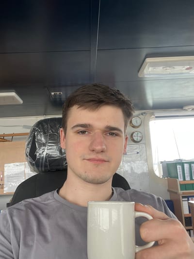 A young adult male, with short brown hair and light facial stubble, wearing a grey t-shirt, is depicted from the chest up, looking directly at the camera. He holds a plain white ceramic mug in his right hand. The scene is located within a ship's bridge in Bangor, United Kingdom. Behind the individual, a black chair with a plastic-covered backrest is visible on the left, adjacent to a wooden rail and documents taped to a bulkhead. To the right, two brass-rimmed gauges are mounted vertically on a light-colored bulkhead, with additional documents below them. A window or port shows a bright, featureless sky or water beyond. Further right, a shelf unit holds several green binders, including ones labeled "CARGO PLANS" and "LOADING BOOK", along with other files. The ceiling is dark blue and paneled, featuring light fixtures.