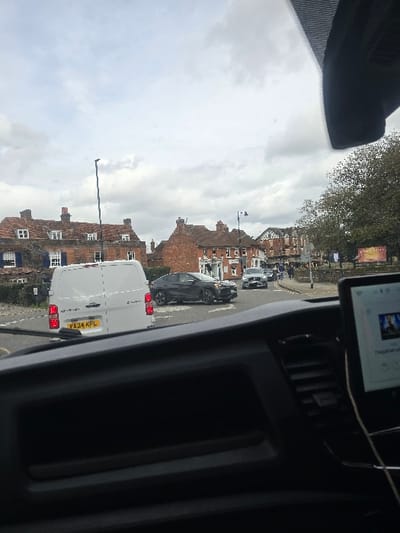 The image presents a forward-facing view from the interior of a vehicle at the junction of Portsmouth Road and High Street/Anyards Road in Cobham, Elmbridge, United Kingdom. The sky above is overcast. The lower portion of the frame displays the dark dashboard of the camera vehicle, with a digital screen mounted on the right console exhibiting media content. A white Chevrolet panel van, bearing UK registration plate `VA24 KPL`, is stationary directly ahead, its rear visible. To the right of the van and slightly beyond, a dark grey SUV is positioned in the roadway. Additional vehicles are visible further down the street on the right. The background features a continuous row of predominantly brick buildings; a distinct structure with blue window shutters is visible on the far left, while a cluster of commercial establishments, including a pub named "The George," occupies the central background. Several pedestrians are discernible walking on the sidewalk to the right. Deciduous trees with green foliage are present on the right side of the street.
