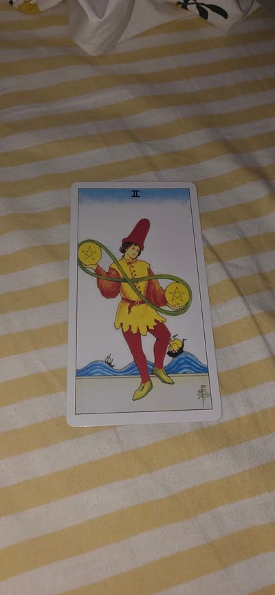A tarot card, the Two of Pentacles, lies on a bed of yellow and white striped fabric. The card depicts a jester-like figure in a red hat and yellow tunic, juggling two gold coins connected by a green ribbon. The background of the card features a blue sky, a blue sea with waves, and two ships. A small, overturned boat is also visible in the water. The fabric underneath the card is textured with diagonal stripes. There is no information about Lieli, Switzerland.