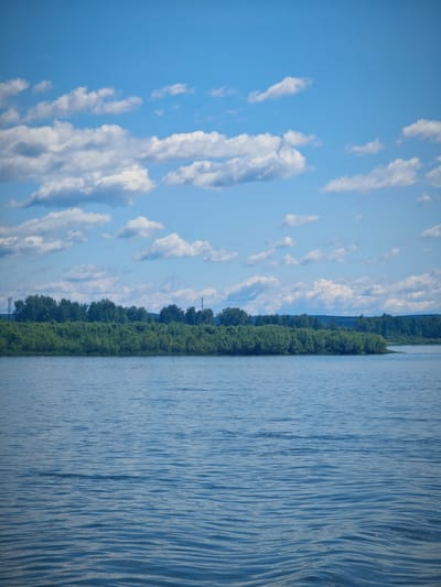 The image shows a wide expanse of calm blue water, with gentle ripples on its surface. In the background, a dense line of green trees forms a verdant bank along the water's edge, suggesting a natural landscape. Above the trees, a bright blue sky is dotted with scattered white clouds, creating a serene and expansive atmosphere. The scene appears to be a natural setting, possibly a river or lake, with no people or man-made structures immediately visible in the foreground or on the tree line, save for two distant, slender communication towers that rise above the foliage on the left and right sides of the horizon. The overall impression is one of tranquility and natural beauty.
