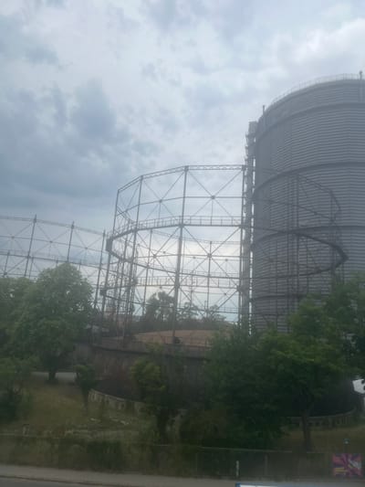 The image shows the historic gasometer in Augsburg, Germany, under a cloudy sky. The tall, cylindrical gasometer, a prominent industrial landmark, is partially obscured by the overcast weather and a complex lattice of metal scaffolding that surrounds it. The scaffolding, with its intricate network of beams and platforms, creates a cage-like structure in front of the gasometer, suggesting a part of its original support system or a more recent construction.

To the left of the main gasometer, another, older section of the structure is visible. This older part appears to be a larger, curved, metal tank, possibly a former gas holder, which is now integrated into the overall site. Trees and greenery surround the base of the gasometers, with some foliage climbing or growing near the structures, indicating a park-like setting or an area that has been reclaimed by nature. The ground around the structures is covered in grass and low-lying vegetation. The overall impression is one of industrial heritage preserved within a natural or semi-natural environment. The scene is static, with no people or active elements visible.