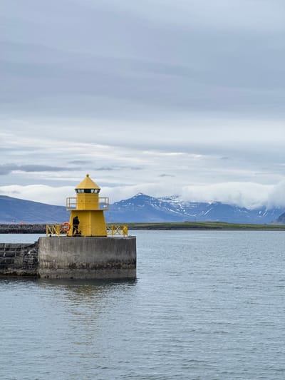 The image depicts a yellow lighthouse situated on a concrete pier in Reykjavík, Iceland. The lighthouse is a bright yellow structure with a square base and a pyramidal roof, surrounded by a yellow railing. A life preserver is attached to the side of the pier. A person is visible inside the lighthouse. The water of the harbor stretches out from the pier, with mountains visible in the background, partially obscured by clouds. The sky is overcast, casting a diffused light over the scene. The pier is constructed from grey concrete and black stones.