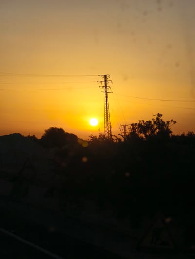 This is a photograph of a sunset in the city of Unknown, Spain. The sun is a bright orb on the horizon, casting a warm, golden light across the sky. In the foreground, silhouetted against the vibrant sky, are several power lines and a tall transmission tower. To the left of the tower, a pile of dark, indistinct material is visible, possibly a construction site or natural debris. Further to the right, a dense line of trees creates a dark, shadowy backdrop. The overall scene is one of natural beauty illuminated by the fading light of day, with man-made structures integrated into the landscape.