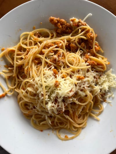 A close-up overhead shot shows a white plate filled with spaghetti and Bolognese sauce, topped with shredded cheese. The pasta is golden brown and appears to be slightly coated in the orange-red sauce. Small pieces of ground meat are visible within the sauce. The cheese is scattered across the top, with some strands melting into the pasta. The plate is clean with some minor smudges of sauce on the rim. The background is out of focus, suggesting an indoor setting, possibly a table in Chipilo de Francisco Javier Mina, Mexico. No people or specific environmental details of Chipilo are visible in the image itself.