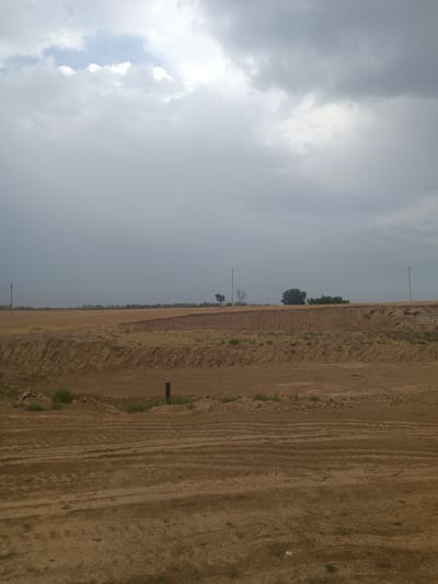 The image depicts a barren, excavated landscape under a cloudy sky. The foreground is a flat expanse of brown dirt with visible tire tracks. In the midground, the land is sculpted into tiered earthen berms, suggesting recent construction or excavation. Some sparse, dry vegetation and rocks are scattered on these berms. A dark, cylindrical object, possibly a marker or post, stands upright in the midground. In the background, a distant line of dry grass and a few scattered trees are visible. Two utility poles with power lines stretch across the horizon. The sky is overcast with a mix of gray and white clouds, with a hint of blue sky visible in the upper left corner. The scene appears to be a construction site or an area undergoing significant land alteration in a rural or semi-rural setting.
