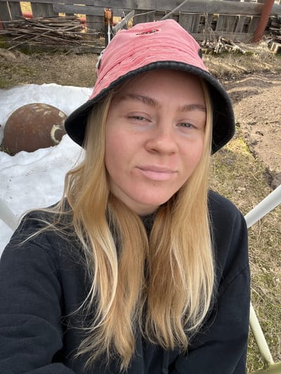 A young woman with blonde hair wearing a black hoodie and a red bucket hat takes a selfie outdoors. The background features a wooden fence, assorted sticks, patches of grass, and a round, brown object resting on a white surface, possibly snow or fabric. The location appears to be a rural or semi-rural area.