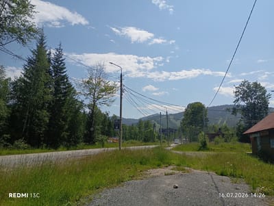 This image depicts a rural roadside scene in the vicinity of Baykalsk, Russia. The viewpoint is from a gravel path leading towards a paved road. On the left side of the road, a dense forest of evergreen trees dominates the landscape, with a few deciduous trees interspersed. A utility pole with numerous wires stretches across the frame, supporting a streetlight. On the right side of the road, a rustic building with a corrugated metal roof is visible, with some signage indicating it might be a shop. In the background, rolling hills covered in trees rise under a bright blue sky with scattered white clouds. A white van is seen driving on the road in the middle distance. The foreground is characterized by tall, green grass and gravel. The image is dated July 16, 2026, at 2:26 PM, as indicated by the timestamp in the lower right corner. The overall atmosphere is one of a quiet, natural environment on a sunny day.