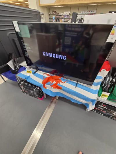 The image shows a large Samsung television on display in a retail store. The television is placed on a table covered with a flag that has blue and white stripes and a red crest with a dragon. Below the table, there are two boxes, one labeled "Makoni EXTRA." A price tag on the table shows "PROMOTION" and "699.-". Another price tag is visible on the right side of the image for 159.-. In the background, shelves are stocked with various products. The environment appears to be a typical electronics section of a supermarket or hypermarket, likely in Käerjeng, Luxembourg, given the presence of the Luxembourgish flag. No individuals are clearly visible in the image.