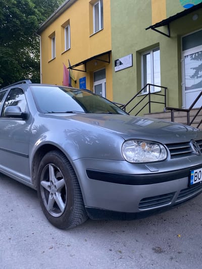 A grey Volkswagen Golf estate car is parked on a paved surface outside a building in the city of Tovstoluh, Ukraine. The building has a yellow facade on the left and a light green facade on the right. Two flags, one pink and blue, and one yellow, are displayed from the yellow section of the building. A plaque with text and a portrait is mounted on the green section of the building. The car's license plate is partially visible and reads "BO". The scene is outdoors during the daytime, with trees visible in the background.