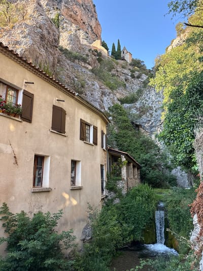 This is a full-frame, eye-level, outdoor photograph taken in Moustiers-Sainte-Marie, France, featuring a residential building nestled beside a steep, rocky cliff face. The building, constructed with a light-colored stucco exterior and a red-tiled roof, has multiple windows, some with brown shutters. Flowers are visible in window boxes. To the right of the building, a smaller stone structure with a covered porch is visible. A small waterfall cascades down a moss-covered rock face into a pool below, surrounded by lush green vegetation and trees. In the background, high up on the cliff, a church with a steeple is perched precariously. The sky is clear and blue. There are no people or animals visible in the frame. The scene conveys a sense of rustic tranquility and integration with the natural landscape.