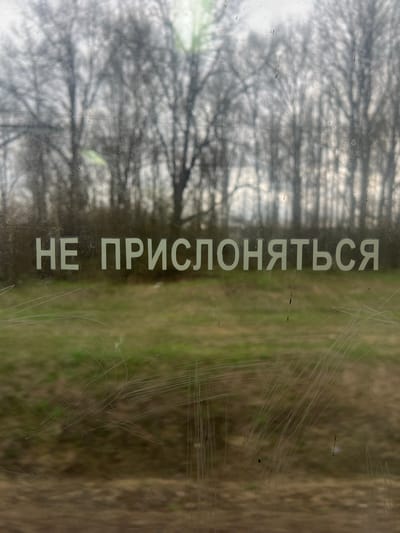 The image displays an outdoor scene viewed through a transparent, heavily scratched surface, likely a window or glass partition, in Nazar'evo, Russia. The most prominent feature is the white Cyrillic text "НЕ ПРИСЛОНЯТЬСЯ" (meaning "DO NOT LEAN" or "DO NOT REST AGAINST") printed vertically on the transparent surface.
The background, seen through the semi-obscured glass, depicts a natural landscape. On the left and center, numerous bare trees form a dense, blurry silhouette against a lighter, possibly overcast, sky. The intricate branches are visible but diffused. To the right, the ground appears as a patch of brownish-green, suggesting grass or dry vegetation.
The transparent surface is significantly marred by numerous fine scratches, particularly concentrated on the right side, which intersect and partially obscure the view. The overall lighting is soft and diffused, characteristic of an overcast day. No people or specific activities are visible. The image captures a static moment, focusing on the warning sign and the muted, natural environment beyond.