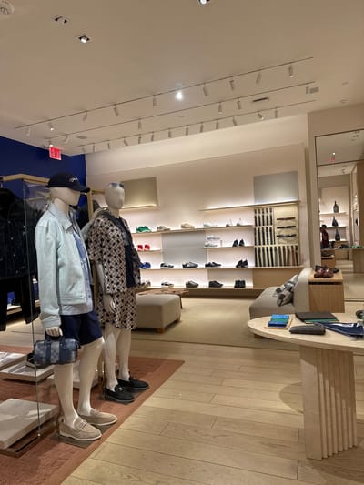 This image captures the interior of a luxurious retail store, likely a high-end fashion boutique. The setting features a sophisticated and well-lit environment, showcasing various apparel and accessories.

In the foreground, two male mannequins are displayed. The mannequin on the left wears a light blue denim-style jacket over a lighter shirt, dark blue shorts, a dark baseball cap with white text (possibly "Louis Vuitton"), and light-colored loafers. It also sports sunglasses and a blue and grey checkered crossbody bag. The mannequin behind it is dressed in a beige and brown checkered pattern shirt, dark trousers, and black loafers.

The store's interior is furnished with light-colored wooden floors and a reddish-brown rug in the display area. The walls are mostly light-colored, with a prominent royal blue section on the left. The ceiling is equipped with numerous track lights and recessed spotlights, providing bright illumination.

Various merchandise is neatly arranged on shelves and display tables in the background. Several pairs of sneakers and shoes in different colors (white, black, red, green) are visible on illuminated shelves. Above them, belts are displayed on horizontal rods. A cream-colored couch and a round, light-colored table are positioned to the right, holding small leather goods like wallets and cardholders, some bearing "LV" or "Louis Vuitton" branding. A blue and white item, possibly a towel or scarf, also shows "Louis Vuitton" text.

Further in the background, mirrors reflect parts of the store. A red "EXIT" sign is visible on a blue wall section on the far left. In one of the cropped images, a person in a reddish-brown top can be seen browsing merchandise in the distance. The overall ambiance is upscale and modern, designed for luxury shopping.