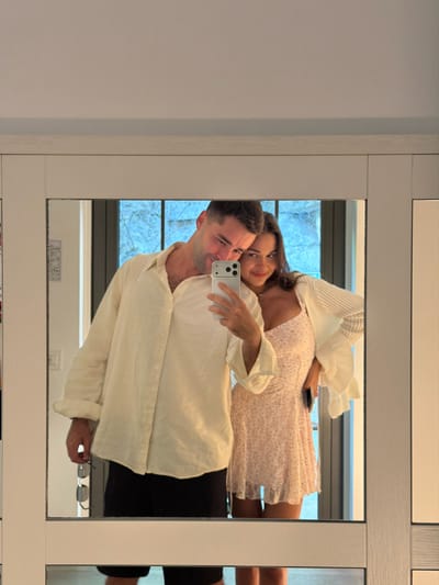 A couple is taking a mirror selfie in a room that appears to be in Pognana Lario, Italy. The man, on the left, is wearing a light-colored, long-sleeved button-down shirt and black shorts. He is holding a pair of sunglasses in his left hand. The woman, on the right, is wearing a light pink floral print mini dress with a white cardigan. She is holding a smartphone in her right hand, capturing the reflection. The background visible through a window in the reflection shows a blue-tinted, possibly stained-glass or textured glass pane. The mirror is framed by a white wooden structure.