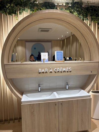 An interior view depicts a circular "Bar à Crèmes" counter within a commercial establishment in Marcq-en-Barœul, France. The central feature is a large, light wood-toned circular unit. The upper half of this unit contains a mirror reflecting the interior space. In the mirror's reflection, a person is visible, holding a light-colored smartphone and seemingly capturing the image. Behind the reflected person, a large, framed artwork with abstract blue and white shapes is visible on a light wood-paneled wall. Also reflected on the counter are two display stands: one featuring a "Google" review QR code and another with text, likely indicating "Lotion hydratante." Several small, gold-colored cylindrical dispensers or decorative elements are present on the reflected counter surface. Above the mirror's reflection, recessed ceiling lights and a vent are visible, along with a patterned ceiling.
On the actual counter unit, below the mirror, the words "BAR À CRÈMES" are spelled out in raised white letters against a light wood panel. Below this, a section of white subway tiles forms a backsplash for a white dual-basin sink unit equipped with two chrome faucets. The lower section of the counter unit consists of two light wood-toned cabinet doors with small, dark handles. Faux green ivy foliage is draped along the top edge of the circular unit and extends onto the adjacent light wood-slatted wall. The floor is light-colored tile. To the right of the counter, a tall, white cylindrical waste receptacle is partially visible.