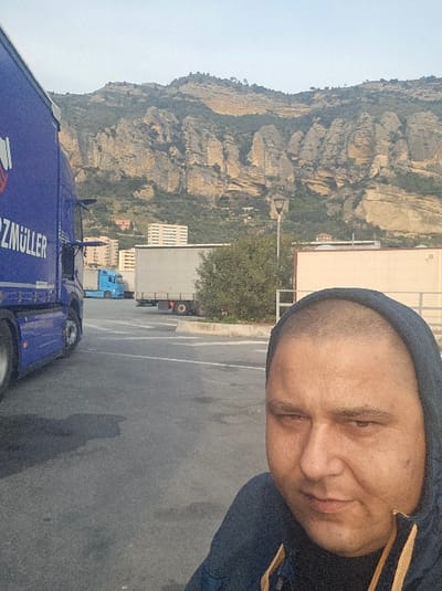 The image captures a selfie of a male individual in a truck parking area located in Ventimiglia, Italy, with a mountainous backdrop.
**Person:** A male subject, appearing to be in his 30s to 50s, occupies the lower right portion of the frame. He has very short or shaved hair and is wearing a dark blue hooded jacket with visible yellow accents near the zipper. His expression is neutral, and his eyes are directed slightly downwards and to the left of the frame.
**Foreground and Midground:** The foreground consists of an asphalt surface, likely a parking lot or truck stop, showing some stains and markings. On the far left, a large blue semi-trailer truck, partially visible, displays the white text "HERZMÜLLER" along its side. Further into the midground, another light blue semi-trailer truck is parked, partially obscured by the foreground truck. To the right of this, a grey semi-trailer is parked behind a low, green bush situated in a concrete island. A dark street lamp post with a single fixture stands vertically in the midground, to the right of the bush. A light-colored, single-story building is partially visible on the far right midground, behind the male subject's shoulder.
**Background:** The background is dominated by a range of rugged, rocky mountains exhibiting light brown and beige rock faces interspersed with green vegetation. At the base and on the lower slopes of these mountains, several buildings are visible, including a prominent multi-story white building with multiple windows. The sky above the mountains is clear and light, indicating daytime.
