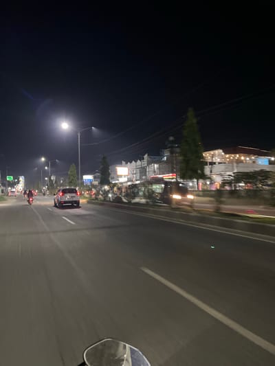 This media file depicts a nighttime scene on a road in Dalam, Indonesia. The perspective suggests the photo was taken from a moving vehicle, possibly a motorcycle given the presence of a side mirror in the lower right corner.

The main subjects are the road, buildings lining one side, and several vehicles. The buildings appear to be shops or businesses, illuminated by interior lights and outdoor signage. One building has decorative string lights. Trees are visible between the road and the buildings. Vehicles on the road include cars and several motorcycles, all with their lights on. One white car is prominently in the middle of the frame, and several motorcycles are clustered further down the road.

The activity is traffic on a road at night. The lighting indicates it is dark, likely late evening or night. The weather appears to be clear. There are streetlights visible, contributing to the overall illumination. There are no distinct people visible, but their presence is implied by the vehicles.

Notable details include the motion blur, indicating movement. The side mirror is reflecting the scene behind the viewer, but it's too blurry to discern details. A faint blue lens flare is visible in the lower left portion of the image. There are some signs visible on the buildings, but the text is unreadable due to the distance and resolution. The overall atmosphere is that of a busy, illuminated road at night in an urban or semi-urban setting.