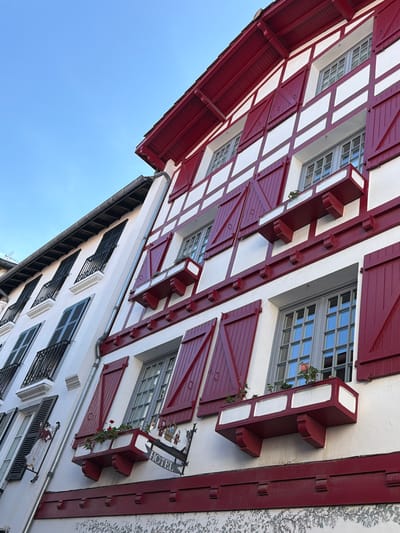 The image displays an upward-angled view of two adjacent multi-story buildings, characteristic of traditional Basque architecture, located in Saint-Jean-de-Luz, France. The primary building on the right features white stucco walls with prominent dark red timber framing and matching bi-fold shutters on its numerous windows. Several windows are adorned with dark red window boxes containing small green plants and red flowers. A wrought-iron sign with "HOTEL" in white lettering and an arrow pointing left, accompanied by ornate figures, is mounted below a window box on a lower floor. A silver metal downspout runs vertically along the seam between the two buildings. The adjacent building on the left has white stucco walls with black bi-fold shutters, some closed and some partially open, and black wrought-iron balconies on its windows. A decorative white and red figurine of a woman in traditional attire is affixed to the wall of the left building. The upper portion of the image shows a clear, bright blue sky.