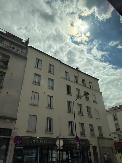 This media file depicts a view of buildings and the sky, taken from an upward-angled perspective. The primary subjects are the facades of residential or commercial buildings, characterized by multiple windows and architectural details such as balconies and external lighting fixtures. On the right side of the image, a vertical sign for "CENTURY 21 L.M.B." is prominently displayed, indicating a real estate agency. Below this, a circular "no parking" sign is visible.
The sky is filled with scattered clouds, suggesting a daytime scene with variable weather conditions. Sunlight breaks through in some areas, creating bright spots and illuminating the clouds. The overall impression is of an urban environment.
While the image is primarily focused on the architecture and sky, a person can be seen through a window in the background, suggesting indoor activity. The scene appears to be set in Alfortville, France, as indicated by the provided location context. The lighting suggests it is daytime, possibly late afternoon or early morning given the angle of the sun. No specific emotions are conveyed, and the activity is primarily static observation of the urban landscape.