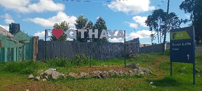The image shows a sign in Othaya, Kenya, located at the coordinates -0.562, 36.941. The sign reads "I LOVE OTHAYA" with a large red heart in the middle. The sign is made of white letters on a white background and is mounted on a metal pole in front of a corrugated metal fence. The fence is partially obscured by green grass and small trees. There is also a KCB bank sign pointing towards a branch and ATM. The sign is located on the side of a dirt road and is surrounded by green grass and some rocks. The background shows a blue sky with white clouds, and some trees are in the background on the right side of the image.