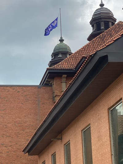 The image shows a view of the rooftop and upper floors of a brick building in Sainte-Anne-de-Bellevue, Canada. A blue flag with white lettering is visible flying from a flagpole atop a green dome. The flag displays text, but it is reversed and partially obscured. The building has a red tile roof and ornate architectural details on the domes. The sky is overcast and gray. The scene is a detailed exterior shot of the building.