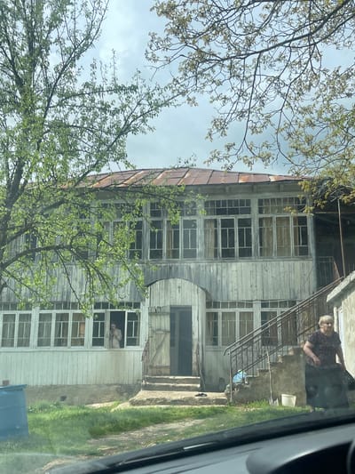 A photograph captures an old, weathered, three-story building with numerous windows, some of which are boarded up, possibly located in or near the city of Chiatura, Georgia. The building is constructed of wood, showing signs of age and disrepair, with a rusty metal roof. The architecture includes a curved section on the left side and an external metal staircase on the right.

An elderly woman stands at the top of the staircase. Another individual is visible in one of the windows on the lower level, observing the scene.

The environment includes foliage, suggesting a rural or semi-rural setting. The view is possibly from inside a vehicle, indicated by part of the car’s interior visible on the right. The overall impression is one of age, abandonment, and a life still lived in the midst of decline.