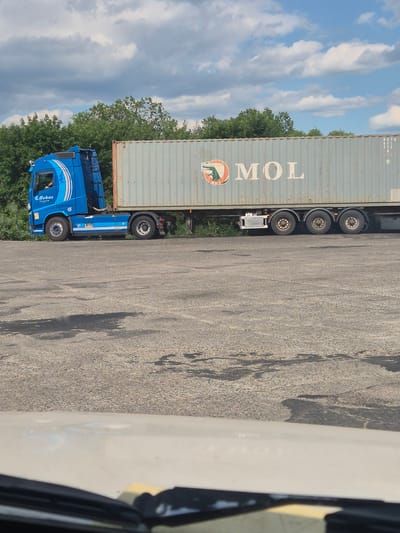 A blue semi-truck with a grey shipping container is parked on an asphalt surface, likely a lot or road, in Pidhaitsi, Ukraine. The truck has the name "Bohns Transport" visible on the side of the cab. The shipping container has the logo and text "MOL" printed on it. Green trees are visible in the background, and a cloudy blue sky is overhead. The asphalt surface shows signs of wear and tear, with some dark patches possibly indicating oil stains. The image appears to be taken from inside another vehicle, as the bottom of the frame shows a portion of a car's interior, including the windshield wiper and dashboard.