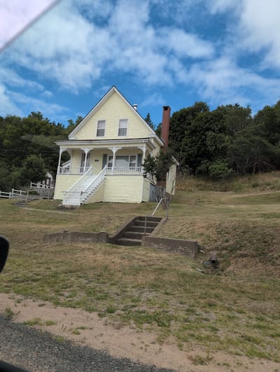 The image shows a yellow, two-story house with white trim, a red brick chimney, and a steep white staircase leading to the front porch. The house is situated on a grassy hillside, with a dense forest in the background. In the foreground, there are concrete steps and a drainage pipe. The sky is blue with scattered white clouds. The image appears to be taken from a moving vehicle, as suggested by the blurred foreground. The location is identified as West Isles Parish, Canada.
