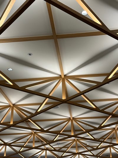 The image displays an upward-facing view of an interior ceiling structure within an unspecified building in Taoyuan City, Taiwan. The ceiling features a complex geometric pattern composed of light-toned wooden beams against a white, flat surface. The wooden beams intersect at multiple points, creating a repeating grid of triangular and star-like formations. Numerous circular recessed light fixtures are embedded within the white ceiling panels. Integrated linear lighting elements are present along some of the wooden beams, casting prominent, radial shadows from the beam intersections onto the white ceiling surface. No individuals, actions, or extraneous objects are visible within the frame.