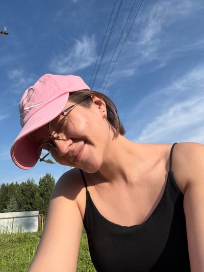 A young person with short brown hair, wearing a pink baseball cap and eyeglasses, looks down with a slight smile. They are wearing a black tank top. The background shows a bright blue sky with wispy clouds and power lines. Below the sky, there are green trees and a fence. In the distance, a small flying object is visible against the sky. The scene appears to be outdoors, possibly in a rural or suburban area.