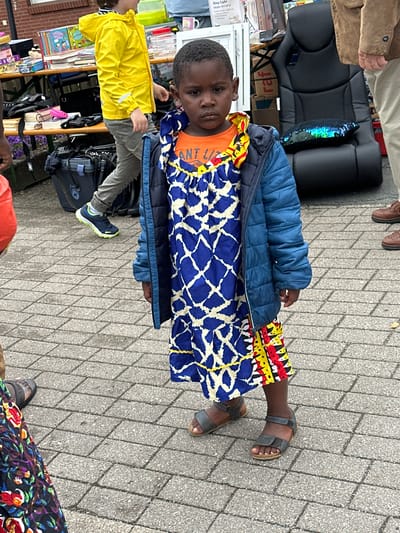This image, taken in Deinste, Germany, depicts an outdoor scene at what appears to be a market or community event, focusing on a young child in the foreground.

The central subject is a young Black child, approximately 4-6 years old, standing still and looking directly at the camera with a neutral expression. The child wears an orange t-shirt, largely obscured by a blue quilted jacket, over a vibrant, multi-patterned dress. The dress features a top section with blue and white geometric patterns, bordered by yellow trim, and a lower section with horizontal bands of red, yellow, and black abstract designs. The child's feet are clad in dark grey/brown sandals with adjustable straps and light-colored soles.

In the mid-ground to the left, a fair-skinned child or youth in a bright yellow hooded raincoat, grey trousers, and blue sneakers with neon green accents is walking from left to right, partially obscured by the foreground. Behind this individual, a wooden table is set up, displaying various items including stacked books and a pink case. Below the table, a large dark travel case with a blue handle is visible.

In the mid-ground to the right, a black ergonomic-style chair is displayed, topped with a shimmering, iridescent sequin cushion that reflects blues, greens, and purples. Partially visible behind the chair are boxes and a white sign with "RING LIGHT" text. To the far right, a light-skinned adult's torso is visible, wearing a brown jacket, with their lower legs in light-colored trousers and brown lace-up shoes. In the far foreground, the lower portion of another person's body is visible on the left, wearing a dark garment with colorful floral patterns and a dark sandal.

The ground is paved with rectangular grey concrete pavers. The lighting suggests an overcast day, producing soft, diffuse shadows. The overall scene indicates an active public gathering or sale.