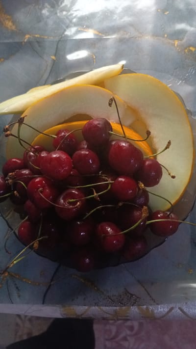 A close-up, high-angle shot captures a bowl filled with dark red cherries and slices of pale yellow melon, presented on a reflective, patterned surface. The cherries, some with stems attached, are piled densely in the foreground. Behind them, curved slices of melon create a backdrop. The surface below appears to be a table with a marble-like pattern, reflecting light and creating streaks of gold and white. The image is taken indoors, likely in a domestic setting in Parenda, Russia, given the context. There are no people or animals visible.