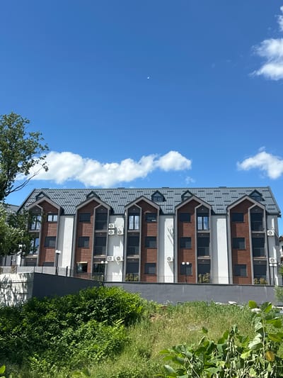 A modern, multi-story apartment building with a dark grey, pitched roof and mansard dormers is situated in Dulovine, Montenegro. The building features white and red-brick facade sections. Numerous windows and balconies are visible, some with dark frames and black railings. Several air conditioning units are mounted on the exterior walls. In front of the building, a grey retaining wall separates it from a dense area of green foliage and grass. To the left, a portion of a mature tree with green leaves extends into the frame. The sky above is a clear, bright blue with scattered white cumulus clouds.
