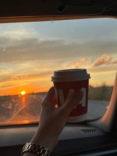 An over-the-shoulder shot captures a hand holding a red disposable coffee cup with a white lid. The cup has white lettering that appears to read "JAVA". The hand is wearing a metallic watch on the wrist. The scene is set inside a vehicle, with the dashboard visible in the foreground. The background, seen through the windshield, shows a sunset with a bright sun low on the horizon. The sky is a gradient of orange, yellow, and blue, with some scattered clouds. The landscape outside appears to be rural, with hints of vegetation. The image has a warm, golden hue due to the sunset lighting.