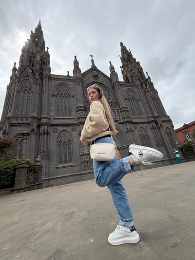 An outdoor, eye-level full-shot captures a young female posing in front of the Parroquia de San Juan Bautista in Arucas, Gran Canaria, Spain. The female, centered in the frame, has long blonde hair and wears a beige teddy-style bomber jacket, light blue baggy jeans, white platform sneakers, and white rectangular sunglasses atop her head. She carries a light-colored "VALENTINO" cross-body bag. Her left leg is raised, bent at the knee, with her foot positioned behind her right calf. She looks directly at the camera over her right shoulder, her lips slightly pursed. The gothic revival church, constructed from dark grey stone, dominates the background, featuring multiple spires, intricate tracery windows, and detailed ornamentation. The sky is overcast with a bright spot visible behind the church's left spire. The ground beneath the subject is a light grey paved plaza. To the left, dark green shrubbery and a black street lamp are partially visible. A reddish-orange building roof is visible on the far right.