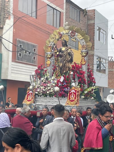 A religious procession is taking place on a street in Callao, Peru. A large, ornate float bearing a statue of Jesus Christ is being carried by a group of men. The float is adorned with numerous red and white flowers, as well as golden embellishments. The statue itself is dressed in a golden robe and is encircled by a large, gilded halo. Several individuals, some wearing traditional religious attire with maroon capes, are participating in the procession, carrying the float and walking alongside it. The background consists of buildings with orange and gray facades, some with visible brickwork and windows. Power lines are prominently visible crisscrossing the sky above the street. A crowd of onlookers is present, some are captured in the foreground and mid-ground, with varying levels of engagement with the procession. One man in the foreground is holding a smartphone and appears to be taking a photograph or video. The sky is overcast.