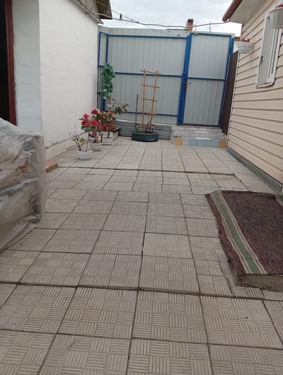 The image shows a paved courtyard in Kostopil, Ukraine. The courtyard is tiled with square, patterned tiles. A white brick building is on the left, and a building with beige siding is on the right. At the far end of the courtyard is a blue-framed gate. Several potted plants are arranged near the gate. A small rug lies on the tiles near the building on the right. An object covered with a plastic sheet is in the foreground on the left. The sky is overcast.
