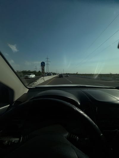 The image shows a view from the driver's seat of a car on a highway. The steering wheel is visible in the foreground, and the dashboard is in the center. The road ahead is clear with several cars on it. The sky is blue with a few clouds. There are power lines and utility poles visible in the distance. The location is identified as Istiqlol, Uzbekistan. The scene depicts a typical highway driving scenario under daylight conditions.