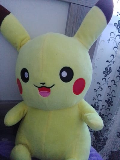 A plush toy resembling Pikachu, a character from the Pokémon franchise, is positioned in the foreground. The toy is primarily yellow with black-tipped ears and red circles on its cheeks. Its eyes are black with white pupils, and its mouth is depicted with a pink tongue sticking out. The toy is seated, with its arms and legs visible.

In the background, a wooden headboard of a bed is partially visible on the left. To the right of the headboard is a window with a curtain. The curtain is white with a dark floral pattern. The lighting in the scene suggests it is indoors.

The provided location details for Nor Geghi, Armenia, are extraneous to the visual content of the image and cannot be substantiated by the image itself.