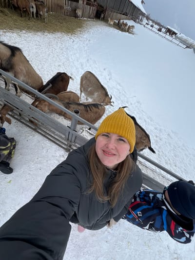 An overhead-angle image depicts an outdoor winter scene at a goat farm in Zaosinovo, Russia. The foreground features a smiling adult female wearing a yellow knit beanie, a dark grey padded winter jacket, and light pink footwear, holding a camera to take a selfie. Her light brown hair is visible beneath the beanie. To her right and partially obscured is a child wearing a dark blue and black patterned winter jacket and a dark blue beanie. Both individuals are positioned on a snow-covered ground adjacent to a metal barrier or fence.
Beyond the barrier, several domestic goats of varying brown, tan, and grey coloration are present on the snow. Some goats are directly adjacent to the barrier, while others are further into the enclosure. In the middle ground, more goats are visible within a fenced-off area with hay on the ground. The background features a rustic wooden barn structure and additional fenced areas, all surrounded by snow. The sky is overcast, indicating a winter day.