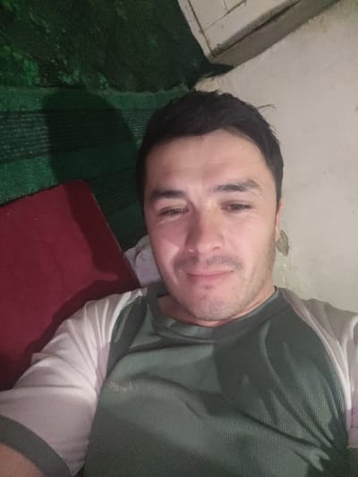 This image depicts a male individual in an indoor setting in Yangikurgan, Uzbekistan. The subject, an adult male with dark hair and light facial stubble, is positioned centrally, looking directly at the camera with a slight smile. He is wearing a two-toned, long-sleeved shirt, consisting of a dark green, ribbed main body and light-colored sleeves. The background to the viewer's left features a dark red object, possibly a cushion or textile, adjacent to a dark green textured surface, likely a mat or fabric. To the viewer's right and behind the subject, a light-colored wall or surface is visible, along with an indistinct white object.