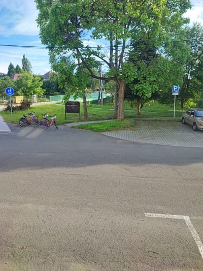 This image depicts a street scene in Nový Jičín, Czechia, bathed in the warm light of late afternoon or early evening. The weather appears to be pleasant, with a clear blue sky dotted with a few clouds. Lush green trees dominate the background, their leaves vibrant and full. A light green bridge spans a waterway in the middle distance, adding a touch of color to the landscape.
In the foreground, a paved area transitions from asphalt to a patterned brick surface. Three pink bicycles with white baskets are parked near a sidewalk, suggesting a recreational or transportation hub. A blue circular sign with white symbols of a pedestrian and a cyclist indicates a shared path, hinting at the area's focus on active mobility. Nearby, a dark wooden information board stands, its contents unreadable.
Further into the scene, a white sign with a blue "P" symbol and an arrow signifies a parking area. A light brown car is parked on the brick surface, partially visible. The overall impression is one of a peaceful, well-maintained urban or semi-urban environment, likely a park or a quiet street designed for both pedestrians and cyclists. The light and greenery suggest a pleasant, summery day.