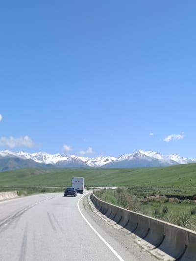 The image depicts a highway with two vehicles on it. A dark-colored sedan is in the left lane, followed by a white semi-trailer truck in the right lane. Both vehicles are traveling away from the viewer. The road is paved and has white lane markings. To the right of the road, there is a concrete barrier. Beyond the road and barrier, there is a vast, rolling green landscape. In the background, majestic snow-capped mountains are visible under a clear blue sky with a few scattered clouds. The overall scene suggests a scenic drive through a mountainous region. The provided context indicates the location is in the city of Ак Жар, Kyrgyzstan.
