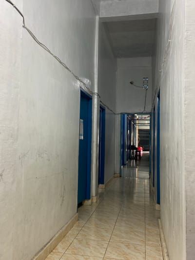 The image depicts a dimly lit, narrow hallway within a building, likely a dormitory or a similar residential facility, located in Tanjung Gading, Indonesia. The hallway is characterized by stark white walls with visible scuff marks and faint graffiti. On the left side, a blue door is visible, and further down the hall, more blue doors line the corridor. The floor is tiled with a beige and brown marbled pattern, reflecting the overhead lighting. Electrical wires are strung along the ceiling and walls, secured by white clips. The hallway extends into the distance, where more rooms or units can be seen, interspersed with patterned flags or banners. In the far background, a red plastic stool is positioned in front of a metal grate. The overall impression is one of functional, somewhat utilitarian accommodation, common in developing regions.