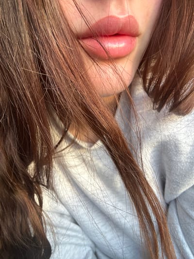 The image presents a forensic close-up view of an individual's lower face, primarily focusing on the mouth area. Full, pink, glossy lips with a prominent highlight on the lower lip are visible at the upper right quadrant, suggesting hydration or lip gloss application. The skin around the lips appears fair. Long, brown-auburn hair, with individual strands discernible, cascades across the frame, obscuring portions of the face and flowing downwards, partially covering the chin and neck area. Beneath the hair and chin, a light-colored, textured garment, resembling a soft, folded sweatshirt, is visible. Warm, natural light illuminates the scene from the right, creating highlights on the lips and hair, and casting subtle shadows. The background is indistinct and out of focus, providing no environmental context. The image does not depict any identifiable geographical location, including Fowlis Easter, United Kingdom. No specific actions or interactions are occurring; the scene captures a static pose.