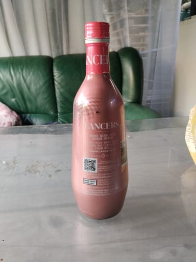 The image shows a pink bottle of Lancers wine, standing on a table. The bottle has a red cap and the "LANCERS" logo is prominently displayed in white text. Several lines of text in different languages are visible on the label, including "VINHO ROSE", "ROSE WINE", "VIN ROSE", "VINO ROSATO", and "VINO ROSADO". There is also a QR code on the label. In the background, a green leather sofa and sheer curtains are visible, suggesting an indoor setting. The lighting appears to be natural, possibly from a window, casting a soft light on the scene. The overall mood is casual and relaxed. The label text indicates that the wine is a rose wine and provides some information about its nutritional content and ingredients, including "Contains: Sulphites".