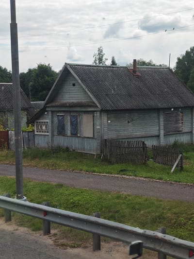 The image captures a weathered, light blue wooden house with a gray corrugated roof in Stukov'ya, Russia. The house appears to be somewhat neglected, with some windows covered by dark plastic sheeting and one window boarded up with plywood. A rusty metal chimney protrudes from the roof. In front of the house, a dilapidated wooden fence encloses a grassy yard. A gravel path runs alongside the property, separated from the foreground by a metal guardrail. The sky above is overcast with scattered clouds. A tall, gray pole, possibly a utility pole, stands to the left of the house. The surrounding area is densely forested with green trees.