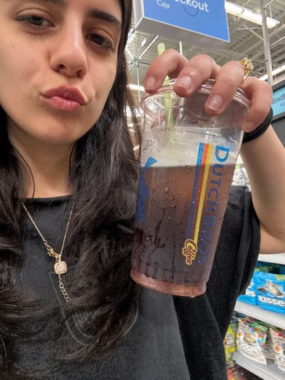 The image depicts a female individual in a selfie-style close-up inside a Walmart retail store, located in Tigard, United States.
The subject, a female with long, dark, wavy hair, is positioned centrally, facing the camera with her lips slightly pursed. She is wearing a black, long-sleeved top and a gold-toned necklace with a square, possibly diamond-set, pendant. Her right hand is visible, holding a clear plastic cup filled with a dark, iced beverage, consistent with coffee or soda. The cup displays the Dutch Bros Coffee logo, featuring vertical blue, red, and yellow stripes and "Coffee" text. Condensation is visible on the exterior of the cup, and a light green straw protrudes from the lid. A large, ornate gold-toned ring with a prominent yellow or amber-colored stone is worn on her right ring finger. Her fingernails are unpolished. A black hair tie is present on her left wrist.
The background shows elements typical of a large retail environment. A blue sign in the upper left corner partially displays "Caja" and "Checkout," indicating a cashier or exit area. To the right, white metal shelving units are stocked with various packaged goods. Blue bags labeled "kisses," consistent with Hershey's Kisses chocolates, are clearly visible on a top shelf, suggesting a candy or snack aisle. Overhead, industrial fluorescent lighting fixtures and structural elements are discernable.